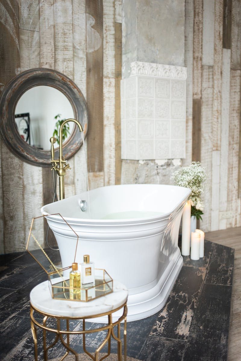 White bathtub near table with glass box of spa supplies placed near wall mirror in modern stylish bathroom with candles